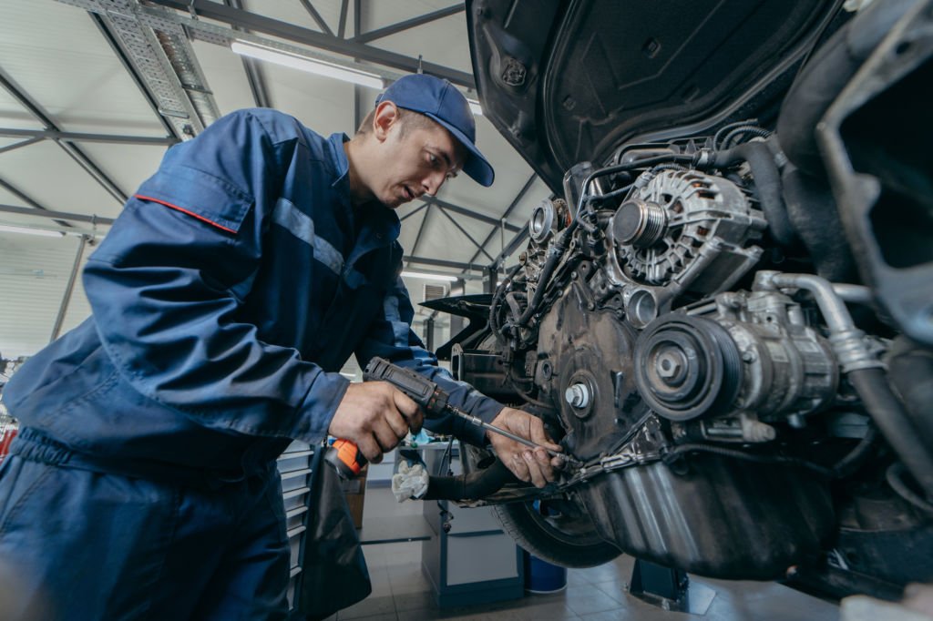 Car mechanic using a cordless impact driver to fix an engine block on a vehicle lift.